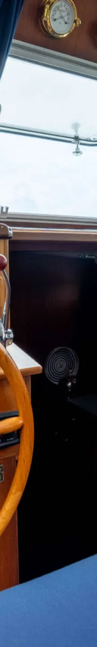 De foto toont het interieur van een stuurhut met een houten stuurwiel, navigatieapparatuur en blauwe kussens.