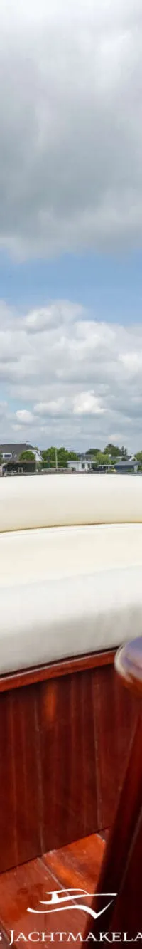 Een boot met crèmekleurige kussens en een houten tafel, varend op een meer met huizen en bomen op de achtergrond.