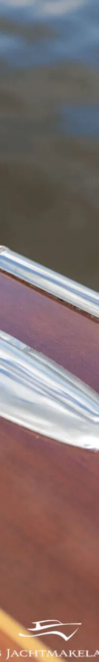 Een detailfoto van een glanzend houten dek met twee chromen beslagen en een logo van Smits Jachtmakelaardij.