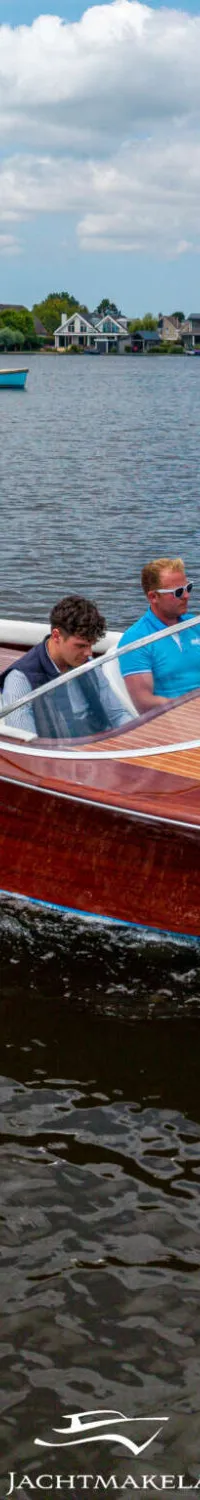 Een houten speedboot met twee mannen aan boord vaart over een meer, met huizen en bomen op de achtergrond.