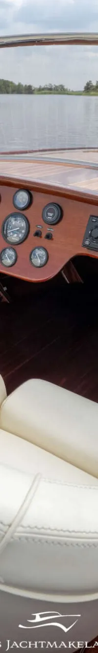 Een houten boot met witte stoelen en een wit stuur, liggend aan een steiger met water en bomen op de achtergrond.