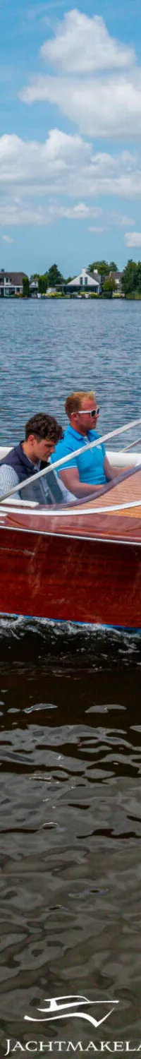 Een houten speedboot met twee mannen aan boord vaart over een meer met huizen aan de oever onder een bewolkte hemel.