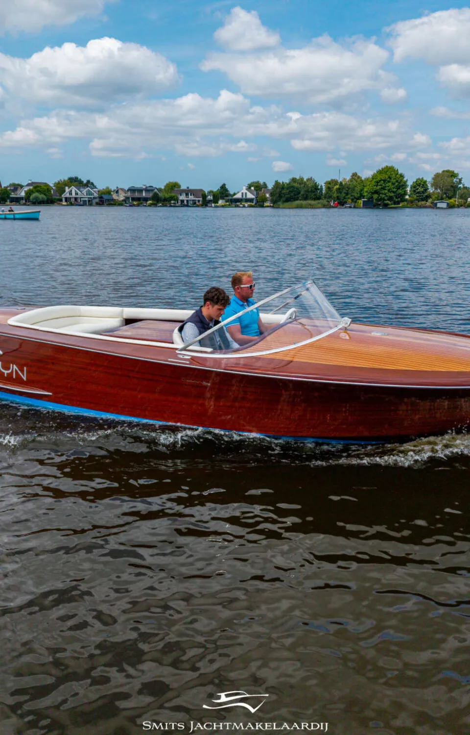 Een houten speedboot met twee mannen aan boord vaart over een meer met huizen aan de oever onder een bewolkte hemel.
