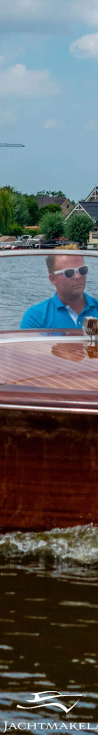 Een houten speedboot met twee mannen aan boord vaart over het water, met huizen en bomen op de achtergrond.