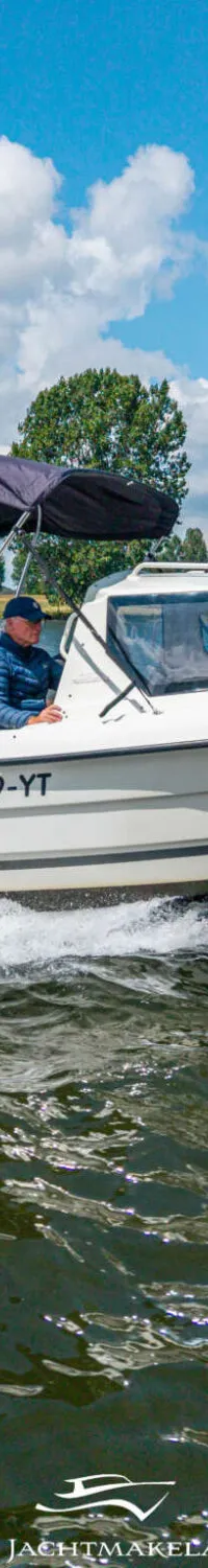Een witte motorboot met een zwarte kap vaart over water, met een man aan het stuur en bomen op de achtergrond.