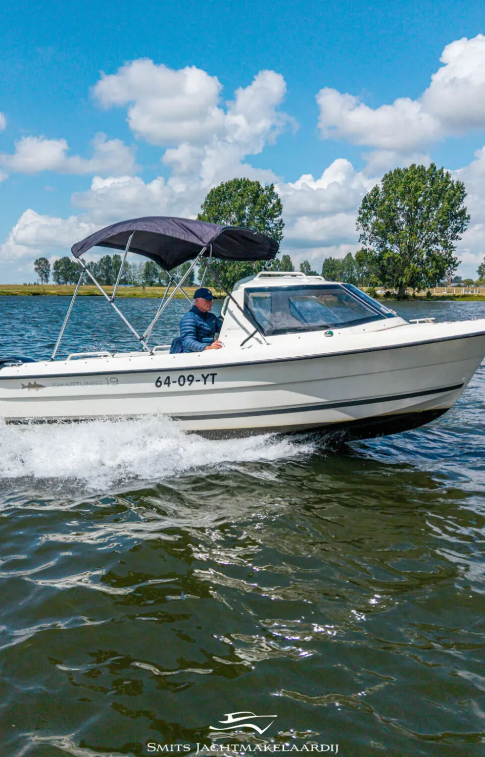 Een witte motorboot met een zwarte kap vaart over water, met een man aan het stuur en bomen op de achtergrond.