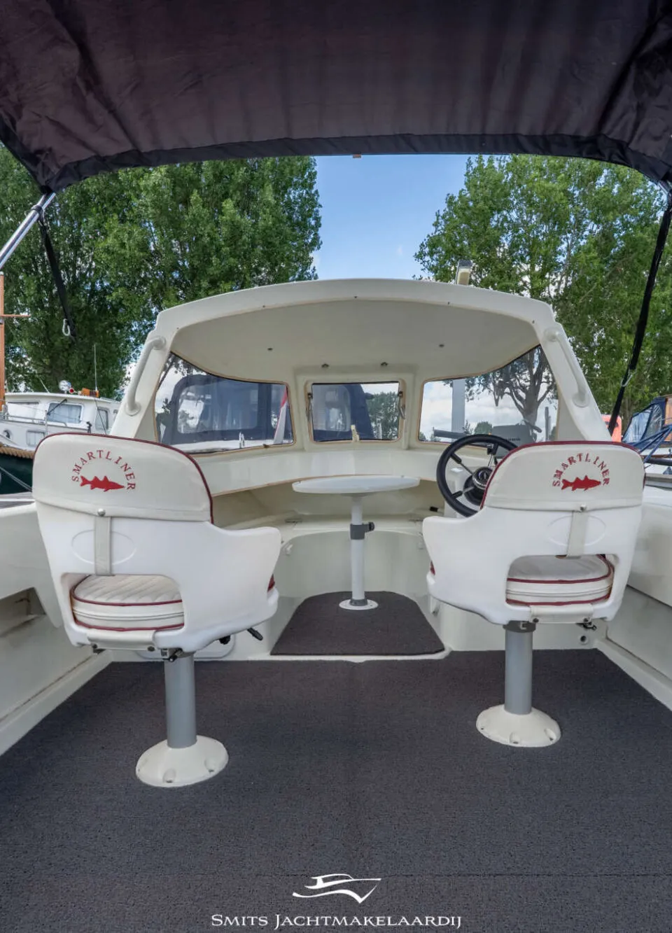 De foto toont het interieur van een boot met twee witte stoelen, een stuurwiel en een kleine tafel.
