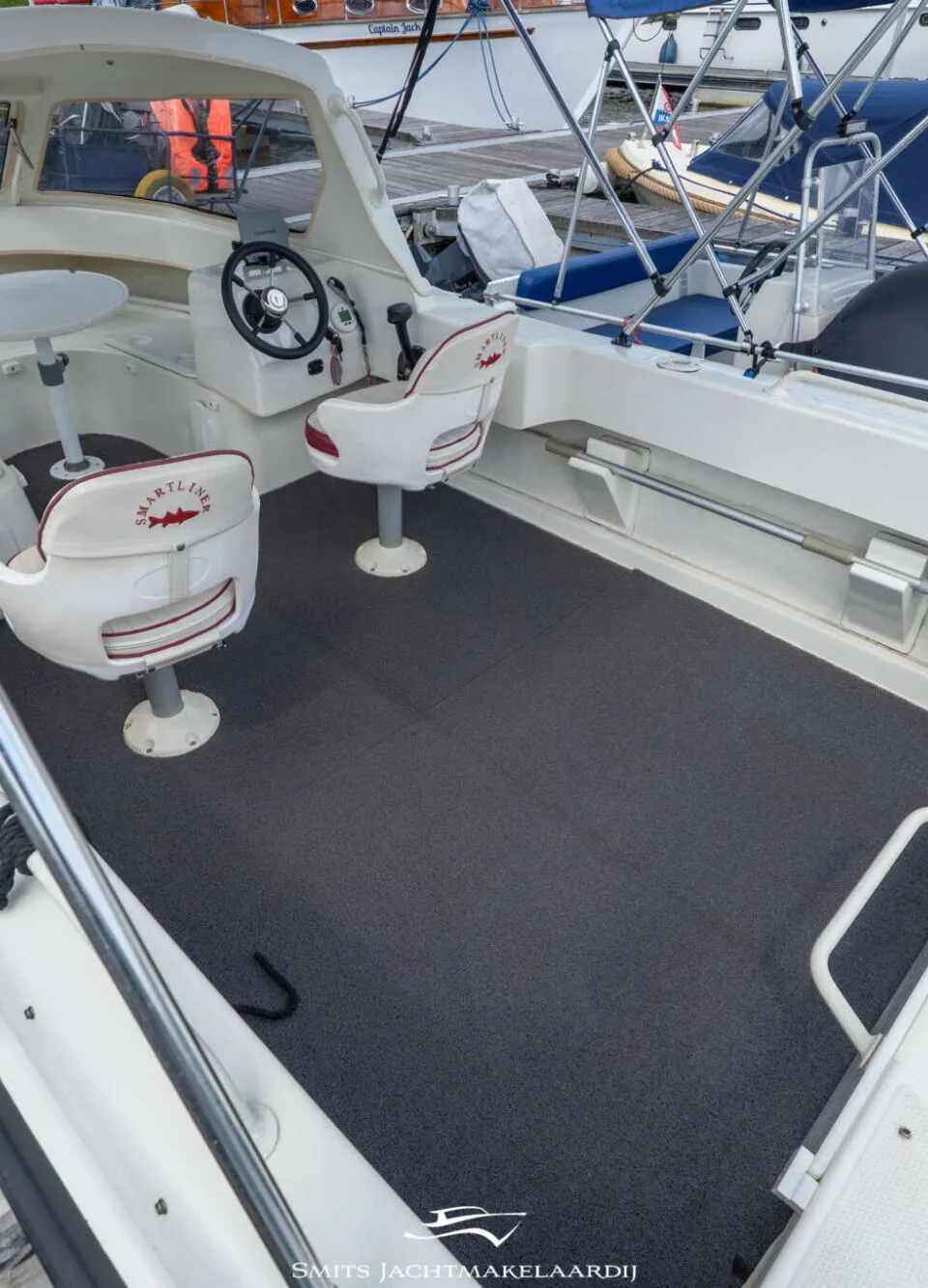 Een witte boot met een donkergrijze vloer, twee witte stoelen, een stuur en een tafel, liggend aan een houten steiger.