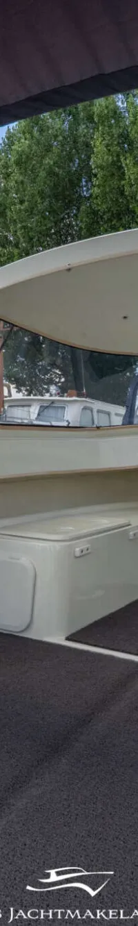 De foto toont het interieur van een boot met twee witte stoelen en een grijs tapijt, met een overkapping en bomen op de…