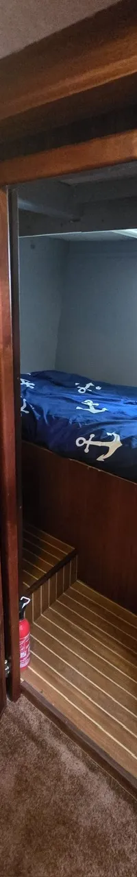 Een open houten deur leidt naar een kajuit met een bed met blauw ankerpatroon beddengoed, een raam en een brandblusser.