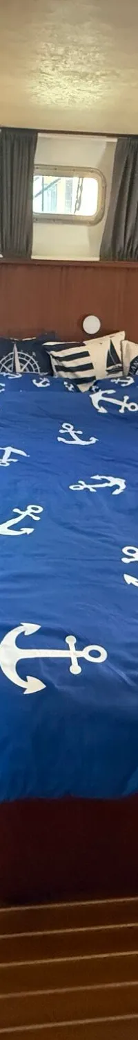 Een slaapkamer in een boot met een bed met blauw dekbedovertrek met ankers, houten wanden en vloer, en ramen met gordijnen.