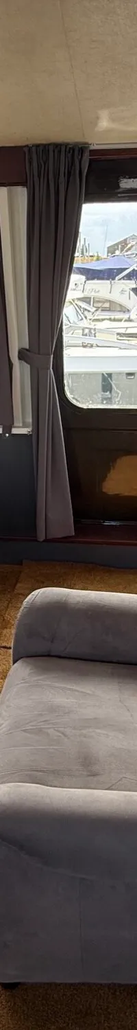 Een interieur van een boot met twee grijze fauteuils, een houten stuurwiel en uitzicht op een haven.