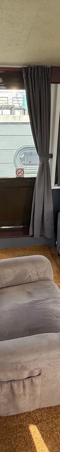 De foto toont een interieur met een bruine vloerbedekking, meerdere grijze fauteuils, een bureaustoel en ramen met gordijnen.