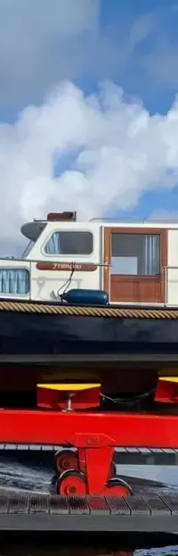 Een donkerblauwe boot met witte opbouw staat op een rode trailer naast een kraan, met een blauwe lucht en wolken.