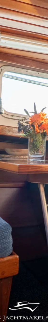 Een interieur van een boot met houten lambrisering, ramen, blauwe kussens en een tafel met bloemen.
