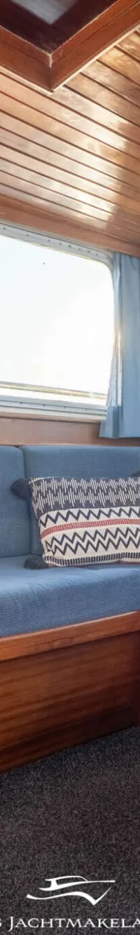 De foto toont een interieur van een boot met houten lambrisering, blauwe banken, ramen met gordijnen en kasten.