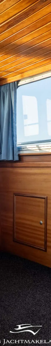De foto toont het interieur van een boot met houten wanden en plafond, blauwe banken en kussens, en ramen met uitzicht op…