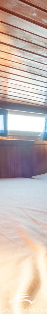 De foto toont een kajuit met houten wanden en plafond, twee bedden met lichte matrassen en ramen met blauwe gordijnen.