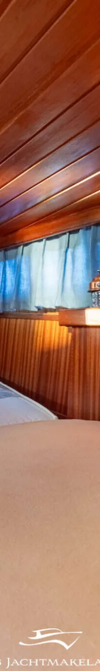 De foto toont een kajuit met houten wanden en plafond, een bed met lichtgekleurd beddengoed en een plank met diverse spullen.