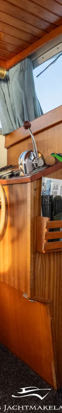 De foto toont het interieur van een boot met een houten stuurwiel, bedieningspaneel en uitzicht op water.
