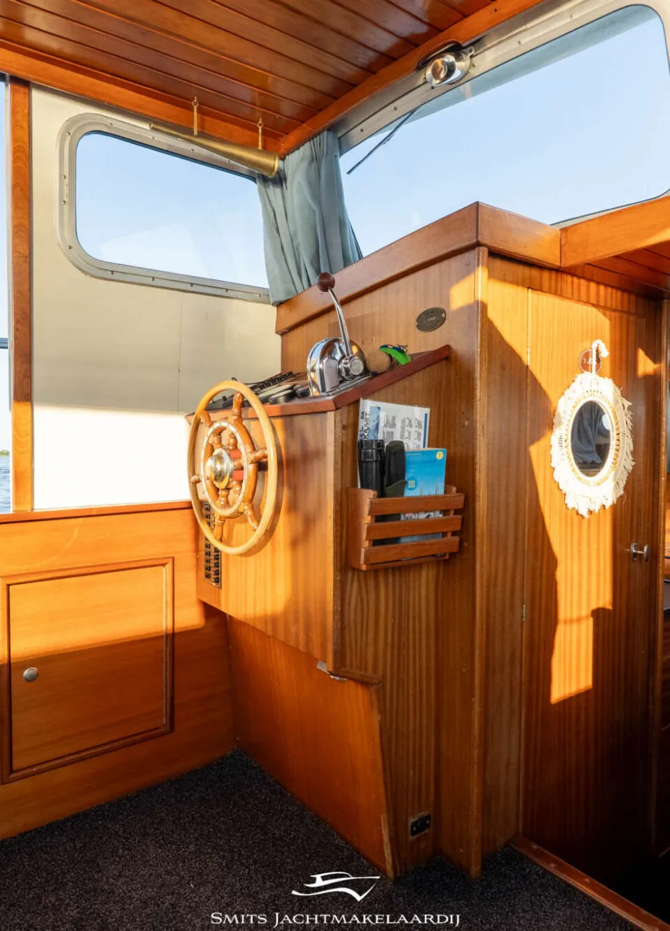 De foto toont het interieur van een boot met een houten stuurwiel, bedieningspaneel en uitzicht op water.