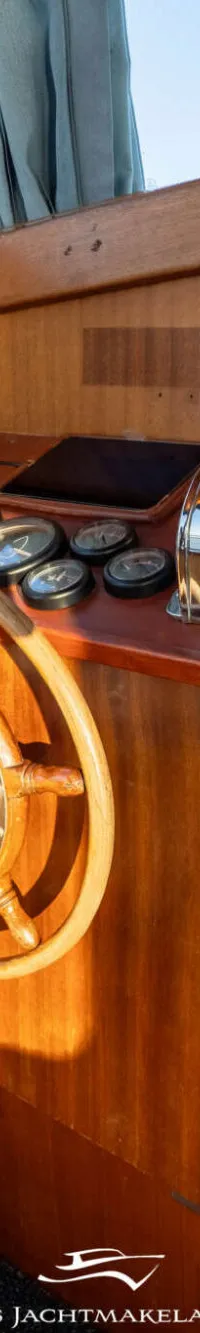 Een houten stuurwiel en bedieningspaneel met diverse meters en een gashendel zijn zichtbaar in de stuurhut.