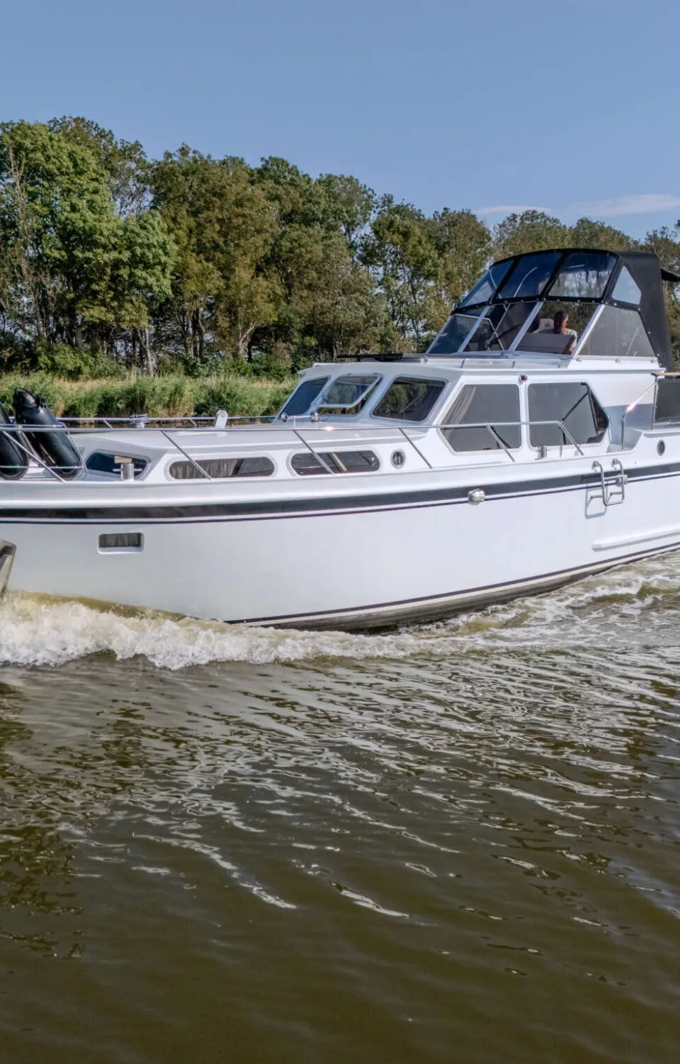 Een witte motorboot met zwarte streep vaart op een waterweg met bomen langs de oever, een persoon is zichtbaar in de…