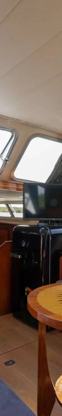 De kajuit van een boot is zichtbaar, met een houten stuurwiel, grijze banken, een tafel met kompasroos en een tv.