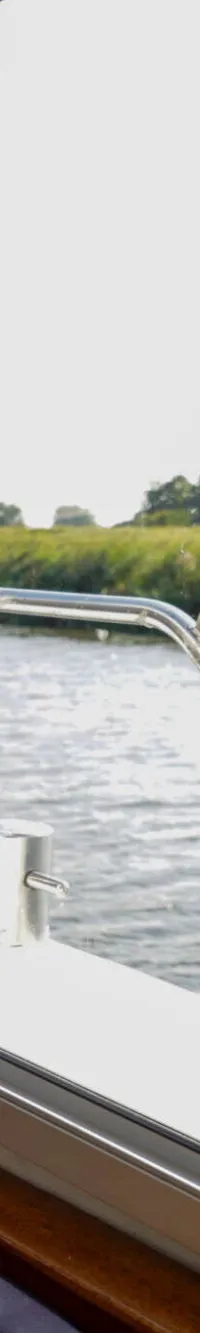Vanuit een bootraam is een waterweg met groene oevers te zien, met een deel van het interieur en de reling van de boot.