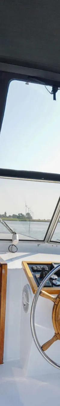 De stuurstand van een boot toont een houten stuurwiel, dashboard, een bankje en de ingang van een kajuit.