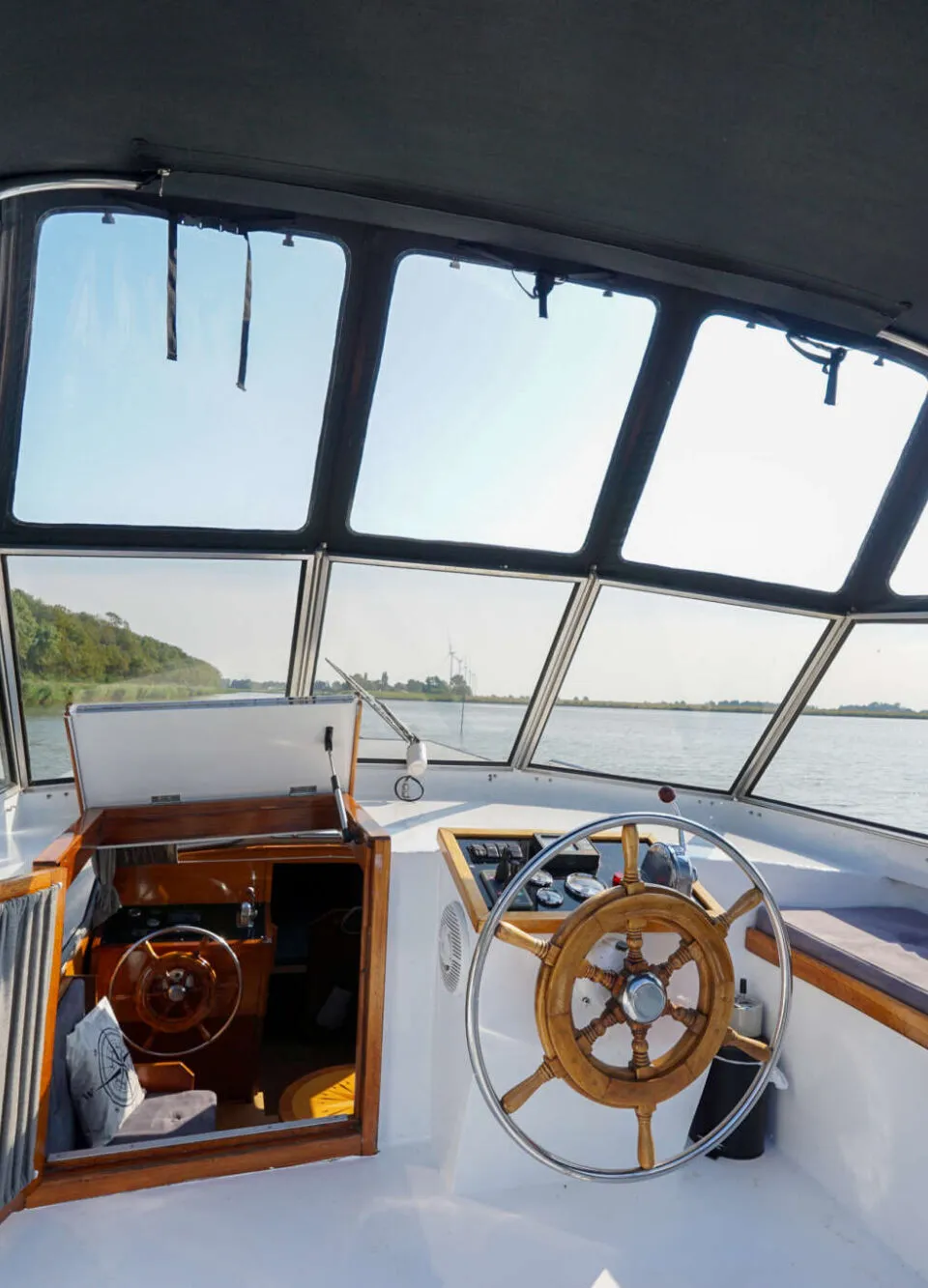 De stuurstand van een boot toont een houten stuurwiel, dashboard, een bankje en de ingang van een kajuit.