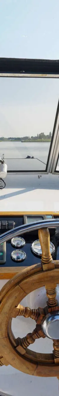 De foto toont de stuurstand van een boot met een houten stuurwiel, instrumentenpaneel en uitzicht over water en landschap.