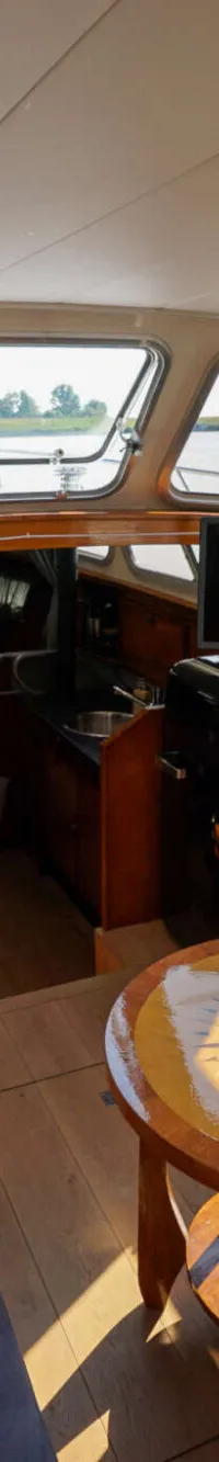Het interieur van een boot met een houten stuurwiel, twee banken, een ronde tafel, een tv en uitzicht naar buiten.