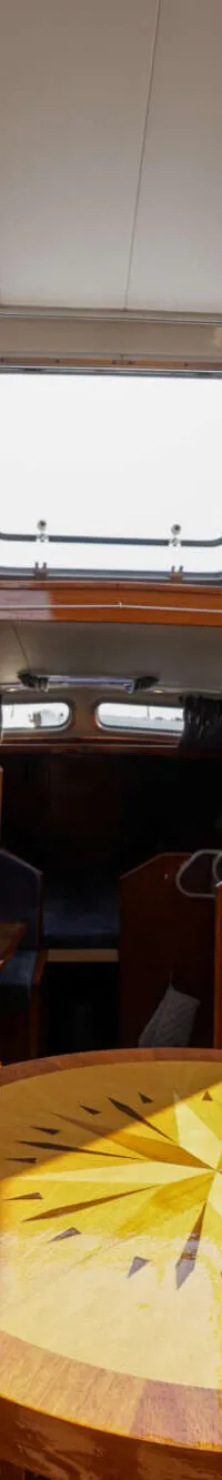 De foto toont een bootinterieur met twee donkergrijze banken, een houten tafel met kompasroos, een stuurwiel en een…
