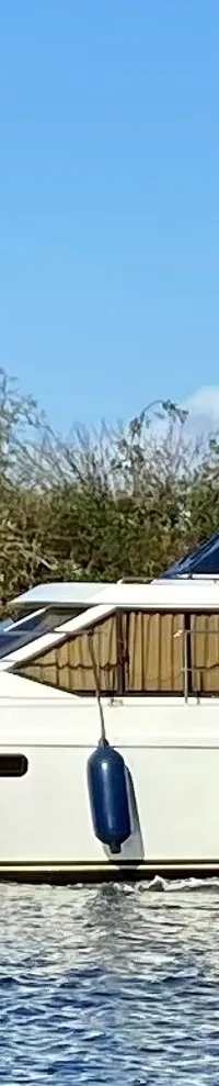 Een witte motorboot met de naam "ANTARIS" vaart op water met een groene oever en blauwe lucht op de achtergrond.