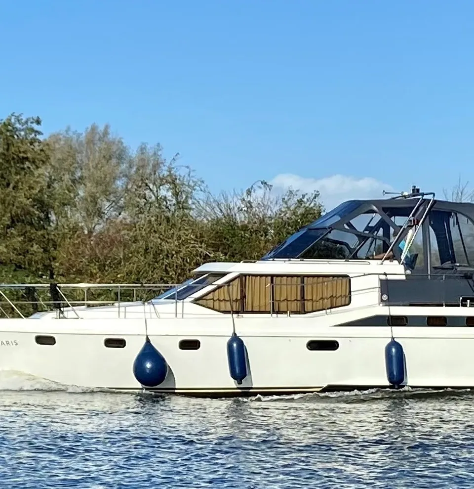 Een witte motorboot met de naam "ANTARIS" vaart op water met een groene oever en blauwe lucht op de achtergrond.