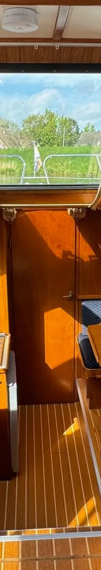 De foto toont het interieur van een boot met houten afwerking, ramen met gordijnen, een bank en een keukenblok.