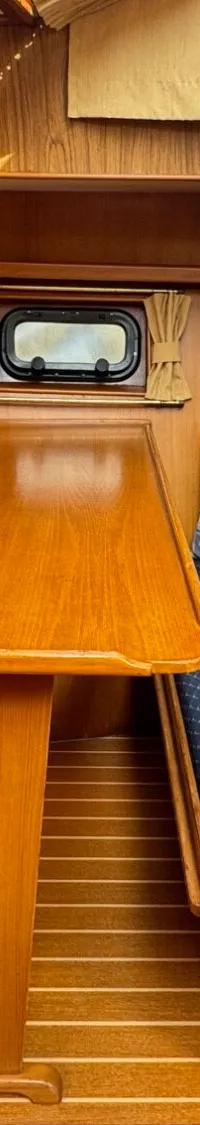 De foto toont een houten interieur van een boot met een tafel, twee banken met blauwe kussens en een patrijspoort.