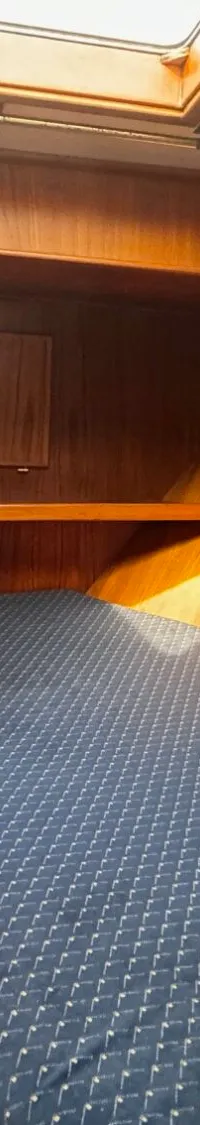 De foto toont een kajuit met een bed met een blauw matras, houten wanden, een dakluik en twee patrijspoorten.