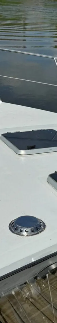 De boeg van een witte boot met reling, twee luiken, een ankerlier en een Nederlandse vlag, varend op water.