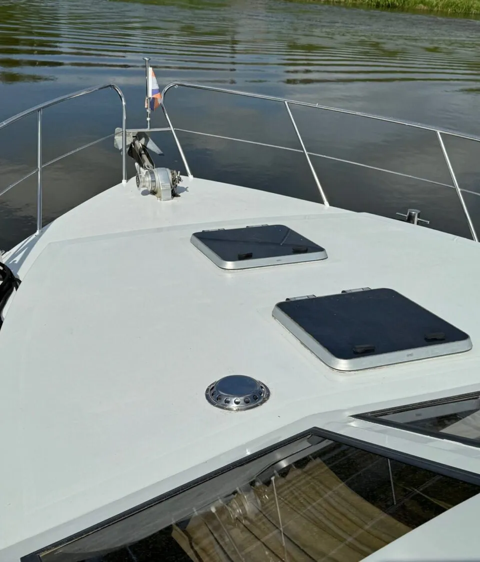De boeg van een witte boot met reling, twee luiken, een ankerlier en een Nederlandse vlag, varend op water.