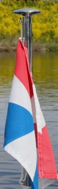 Een Nederlandse vlag wappert aan een vlaggenstok tussen twee metalen relingen, met water en een gele oever op de achtergrond.