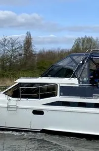 Een witte motorboot met een donkergrijze opbouw vaart op een waterweg met rietkragen en bomen op de achtergrond.