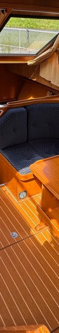 De foto toont het interieur van een boot met houten wanden, vloer en meubilair, blauwe zitkussens en ramen met gordijnen.