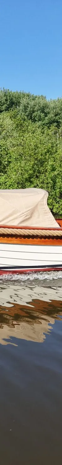 Een man vaart in een witte sloep met houten details en een beige kap, met een Nederlandse vlag achterop.