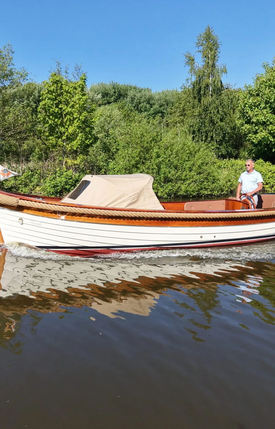 Een man vaart in een witte sloep met houten details en een beige kap, met een Nederlandse vlag achterop.