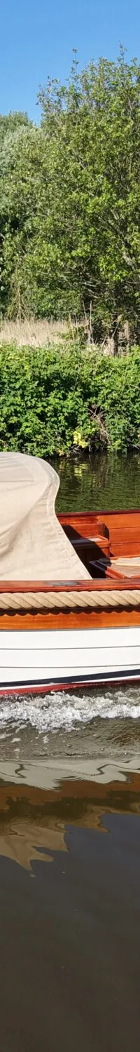 Een man vaart in een witte sloep met houten details en een beige kap op een groene rivier, omringd door bomen.