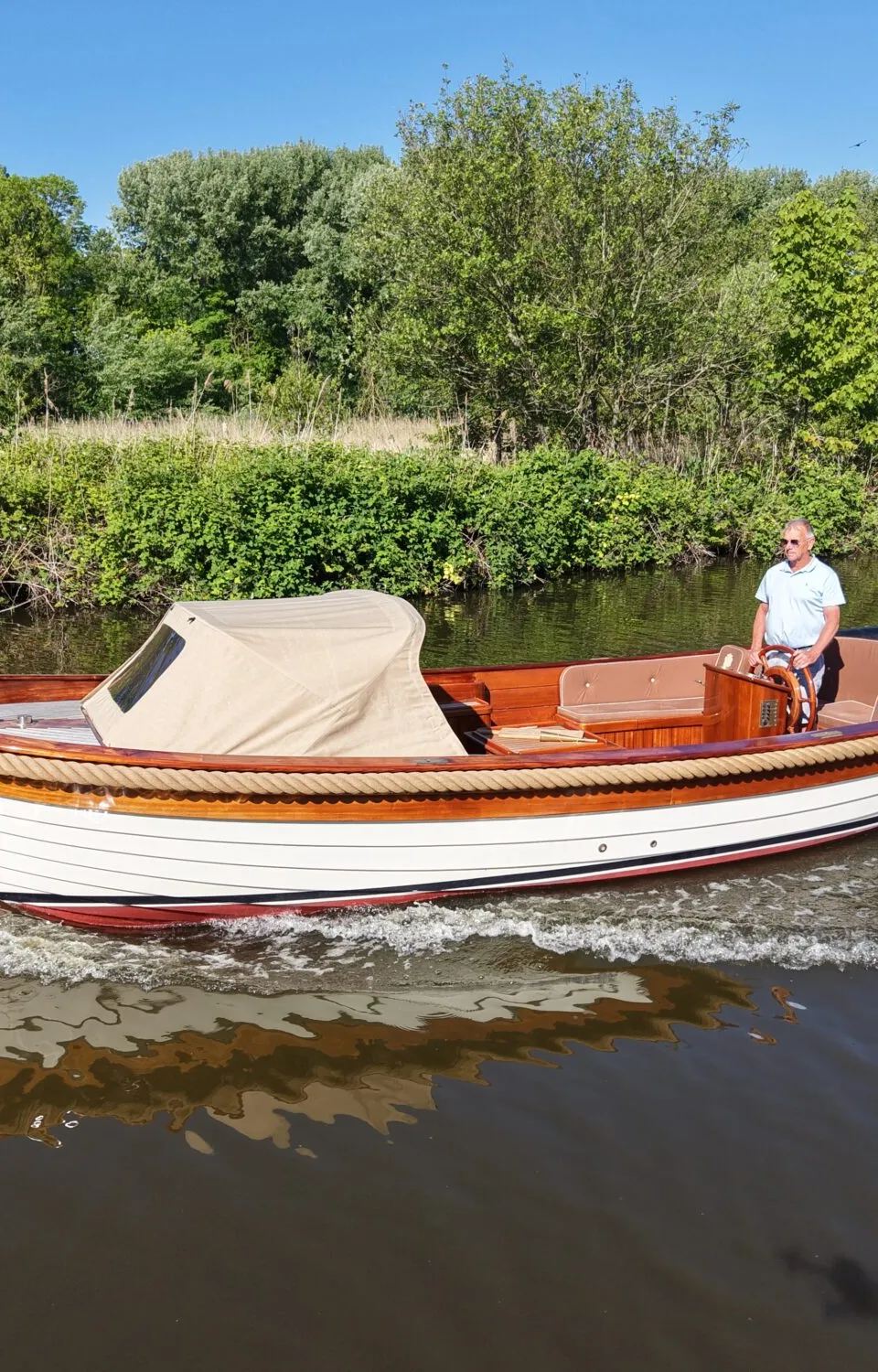 Een man vaart in een witte sloep met houten details en een beige kap op een groene rivier, omringd door bomen.