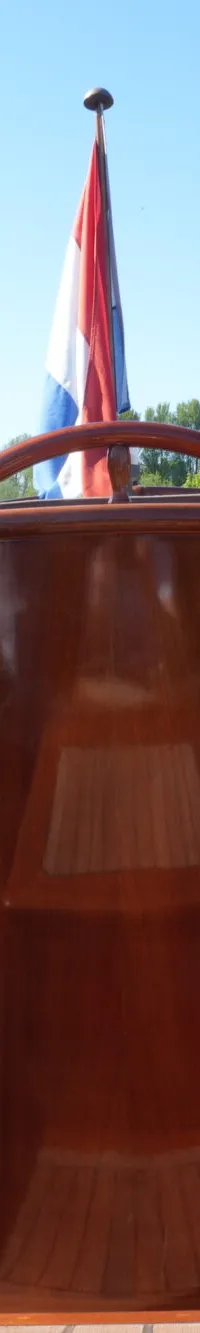 Een houten boot met bruine kussens en een Nederlandse vlag, varend op een waterweg omringd door groen.