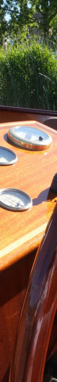 Een houten stuurwiel en dashboard met meters zijn zichtbaar in een boot, met riet en gebouwen op de achtergrond.